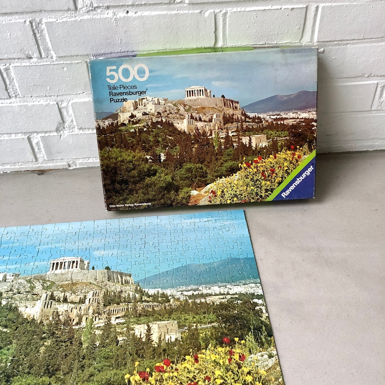 K&ouml;nnte beinhalten: Ein 500-teiliges Ravensburger Puzzle mit einem malerischen Blick auf die Akropolis in Athen, Griechenland. Die Schachtel des Puzzles zeigt ein farbenfrohes Bild der Akropolis mit &uuml;ppigen gr&uuml;nen B&auml;umen und leuchtenden Blumen im Vordergrund.