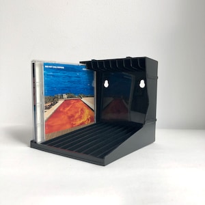 May include: Black CD storage rack with a CD case featuring the Red Hot Chili Peppers album "Californication" with a pool and a red sky on the cover art.
