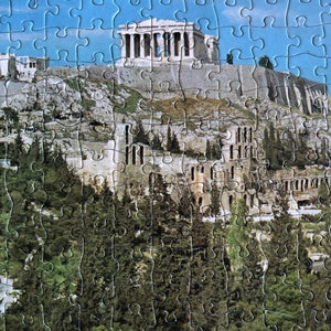 K&ouml;nnte beinhalten: Ein Puzzle mit einem malerischen Blick auf die Akropolis in Athen, Griechenland. Die Puzzleteile sind so zusammengesetzt, dass der ikonische Parthenon-Tempel auf einem H&uuml;gel zu sehen ist, umgeben von B&auml;umen und Geb&auml;uden. Der Himmel ist blau und klar.