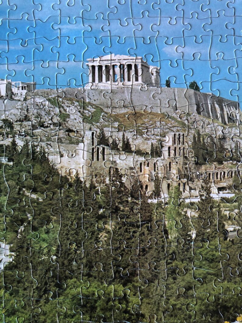 K&ouml;nnte beinhalten: Ein Puzzle mit einem malerischen Blick auf die Akropolis in Athen, Griechenland. Die Puzzleteile sind so zusammengesetzt, dass der ikonische Parthenon-Tempel auf einem H&uuml;gel zu sehen ist, umgeben von B&auml;umen und Geb&auml;uden. Der Himmel ist blau und klar.