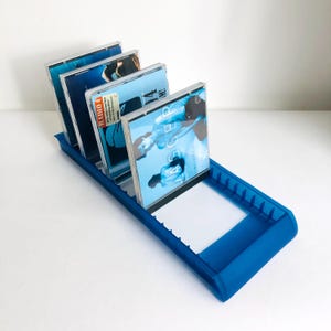 May include: A blue plastic CD holder with four CDs in jewel cases. The CDs have blue and white album art. The holder is rectangular with a raised edge to keep the CDs in place. The holder is on a white surface.