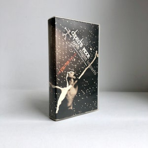 Peut inclure: Une cassette VHS noire avec une image en noir et blanc d'une personne les bras levés. Le texte sur la cassette indique "Depeche Mode one night in Paris" et "The Exciter Tour 2001".