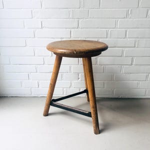 Könnte beinhalten: Ein Vintage-Hocker aus Holz mit rundem Sitz und drei angewinkelten Beinen. Der Hocker besteht aus hellbraunem Holz mit sichtbarer Maserung und Abnutzung. Eine schwarze Metallstützstange verbindet die Beine. Der Hocker ist etwa 46 cm hoch.