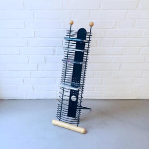 Può includere: Porta CD in metallo nero dal design unico. La rastrelliera presenta una serie di ripiani orizzontali in filo metallico per contenere i CD. Ha una base in legno e due sfere di legno nella parte superiore. La rastrelliera è leggermente inclinata verso sinistra.