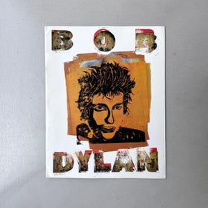May include: A rectangular poster featuring a high-contrast black and orange portrait of Bob Dylan centered on a white background. The name BOB DYLAN is printed in large, distressed, textured block letters above and below the portrait.