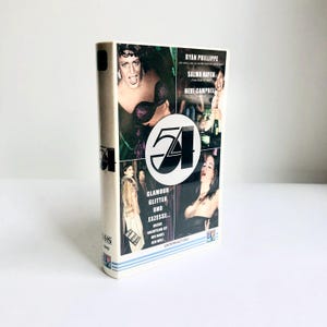 Könnte beinhalten: Eine VHS-Kassette für den Film "Studio 54" mit weißem Gehäuse. Das Cover zeigt Bilder von Schauspielern und den Text "GLAMOUR GLITTER UND EXZESSE..." und "STUDIO 54". Ein Vintage-Unterhaltungsobjekt.