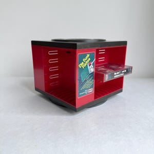 Könnte beinhalten: Ein roter und schwarzer, drehbarer Kassetten-Displayständer. Das Display hat mehrere Schlitze für Kassetten und ein Etikett mit der Aufschrift "Die Funk Füchse". Eine Kassette ist in einem der Schlitze sichtbar.