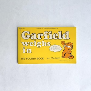 Könnte beinhalten: Ein Vintage-Garfield-Buch mit dem Titel "Garfield Weighs In" auf gelbem Hintergrund. Das Cover zeigt die Cartoon-Katze Garfield mit dem Text "I'm not overweight, I'm undertall." Das Buch ist von Jim Davis.