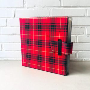 Puede incluir: Un archivador a cuadros rojos con cierre de correa negra. El archivador tiene un patrón de tartán clásico en rojo, negro y dorado. El archivador está de pie sobre una superficie blanca.