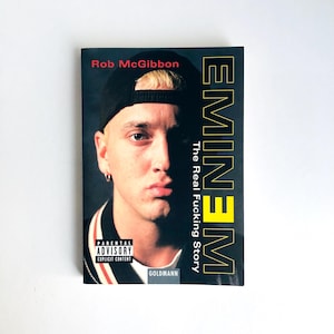 May include: Book cover with a portrait of Eminem in a black cap. The title reads "Eminem: The Real Fucking Story" by Rob McGibbon. The cover includes a parental advisory warning. The book is published by Goldmann.