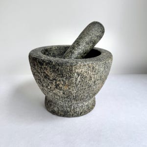 Op de afbeelding: Een zware vijzel en stamper van donkergrijs graniet met een gespikkelde natuurstenen textuur. De vijzel heeft een brede, ronde kom met een dikke rand en een stevige, uitlopende voet, terwijl de cilindrische stamper in de kom rust.