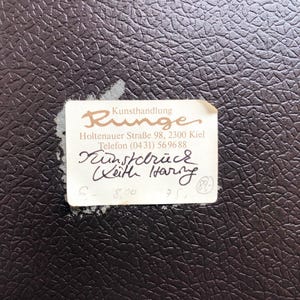 May include: A close-up of a dark brown textured surface with a torn white label. The label features the word "Runge" in a stylized font, along with handwritten text and numbers. The label is slightly torn.