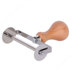 Puede incluir: Un cortador de pasta de acero inoxidable y madera con dos cuchillas circulares. El mango es de madera clara y las cuchillas están conectadas por un marco de metal. El cortador está diseñado para hacer pasta.