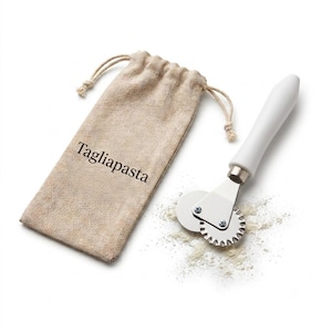 May include: A pasta cutter with a white handle and a dual-wheel design featuring one smooth and one fluted metal blade. Beside it lies a beige drawstring pouch with the word Tagliapasta printed in black, accompanied by a small dusting of flour.