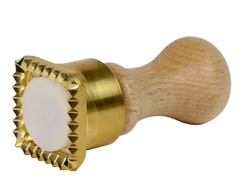 Puede incluir: Un sello para repostería de metal dorado con un mango de madera. El sello tiene un diseño cuadrado con un patrón en relieve de triángulos alrededor de los bordes.