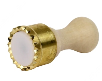 Puede incluir: Un cortador de masa de latón y madera con un borde festoneado y un centro de plástico blanco. El cortador tiene un mango de madera.