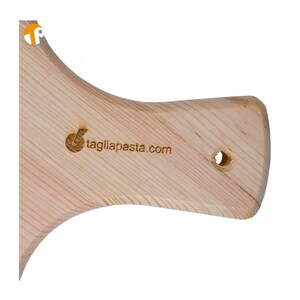 May include: A light-colored wooden pasta cutting board with a handle. The handle has a hole for hanging and the text "tagliapasta.com" is visible. The wood grain is visible.
