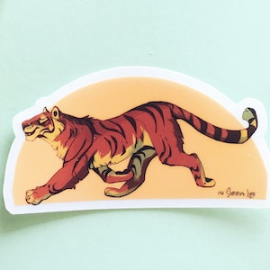 Puede incluir: Una pegatina de un tigre corriendo sobre un fondo amarillo. El tigre es naranja y negro con rayas blancas. La pegatina tiene forma de semicírculo y lleva la firma del artista en la esquina inferior derecha.