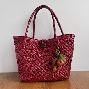 May include: A deep red woven tote bag featuring a textured, open-lattice design, two matching braided handles, and a single dark brown button closure. A decorative charm consisting of small wooden fruit and leaf shapes hangs from one handle.