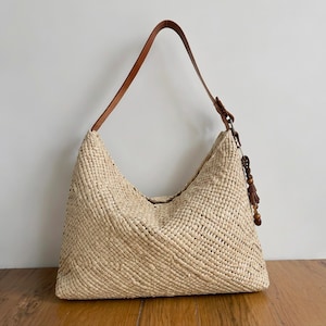 May include: A hobo-style shoulder bag crafted from woven light beige straw material. The bag features a single tan leather strap and a decorative side accent consisting of brown wooden beads and a small tassel hanging from a metal ring attachment.