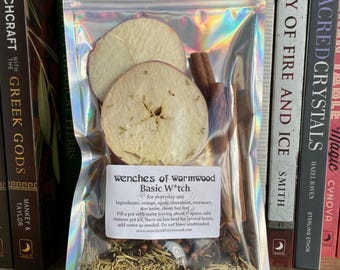 May include: A clear plastic bag filled with dried fruit, herbs, and spices. The bag is labeled "wenches of wormwood Basic W*tch for everyday use". The ingredients listed are orange, apple, cinnamon, rosemary, star anise, clove, and bay leaf.