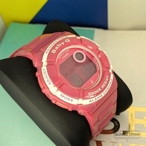 Op de afbeelding: Een roze Baby-G digitale horloge met witte accenten. Het horloge geeft de tijd weer en heeft knoppen voor aanpassing, modusselectie en alarmfuncties. Het is water- en schokbestendig.