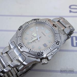 May include: A silver-tone Casio analog wristwatch with a metallic link band. The watch features a light-colored dial with luminous hour markers, a date window at the three o'clock position, and a rotating bezel with black numerals.