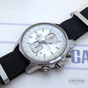 May include: A silver-toned chronograph wristwatch featuring a white dial with three sub-dials, a date window, and a tachymeter scale on the outer rim. The watch has a black nylon strap with silver-toned metal hardware and the text WIRED on the dial.