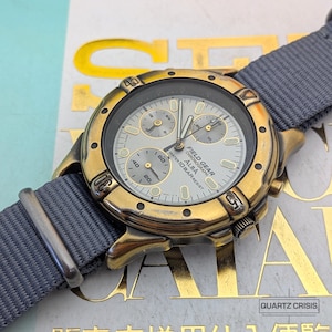 May include: A gold-toned ALBA Field Gear chronograph watch with a grey nylon strap. The watch face is silver with black and gold accents, featuring multiple sub-dials and the text "WATER 10 BAR RESIST". The watch is displayed on a gold and white surface.