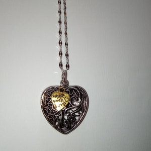 May include: A silver chain necklace with a silver and gold heart pendant. The heart pendant has a filigree design and a smaller gold heart charm with the words "made with love" inscribed on it.