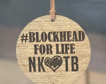 May include: A wooden round ornament with a brown leather cord. The ornament has the text "#BLOCKHEAD FOR LIFE NK❤TB" engraved on it. A small wooden charm with two hearts and an infinity symbol hangs from the ornament.