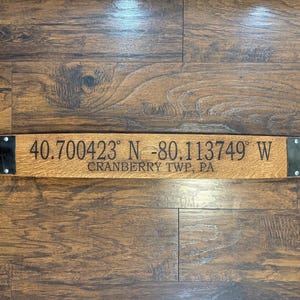 May include: A rectangular wooden sign with the coordinates 40.700423° N -80.113749° W and the text CRANBERRY TWP, PA. The sign is made of light brown wood with black metal accents on each end.