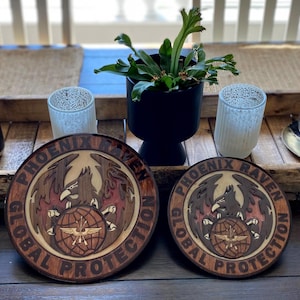 May include: Two wooden round plaques with a brown, red, and white design. The plaques feature a phoenix with the words "Phoenix Raven Global Protection" around the outside.