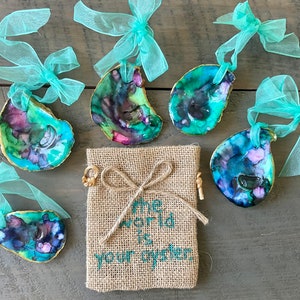 May include: Five iridescent blue, green, and purple oyster shell ornaments with gold trim and turquoise ribbon. The ornaments are arranged around a burlap bag with the text "The world is your oyster" embroidered in green.