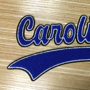 以下が含まれることがあります： 「Carolina」という単語が筆記体で刺繍された青と白の刺繍パッチ。パッチは野球帽の形をしています。