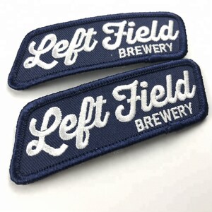 May include: Two navy blue embroidered patches with white stitching. The patches are rectangular and curved at the edges. The text on the patches reads "Left Field Brewery".