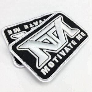 May include: Two white embroidered patches with black borders and white stitching. The patches are rectangular and feature a stylized "M" with the text "MOTIVATE ME" below it.