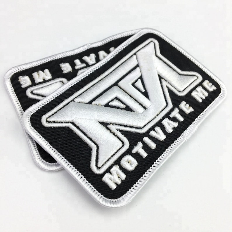 May include: Two white embroidered patches with black borders and white stitching. The patches are rectangular and feature a stylized "M" with the text "MOTIVATE ME" below it.