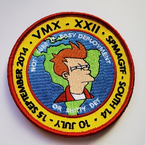 May include: A yellow and red embroidered patch with a cartoon character and the text "VMX - XXII - SPMAGTF - SOUTH 14 - 10 JULY - 15 SEPTEMBER NOT EASY DEPLOYMENT OR SH*TTY DET"