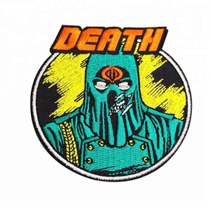 May include: An embroidered patch featuring a green and black character with a yellow and orange background. The character has a skull-like face with a single eye and a Cobra symbol on its forehead. The word "DEATH" is written in orange above the character.
