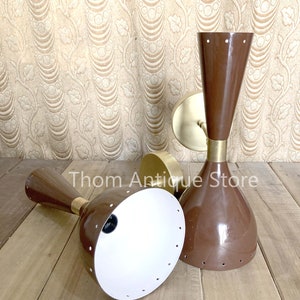 May include: Two brown mid-century modern sconces with brass accents. The sconces have a conical shape with a white interior and small circular perforations. The sconces are on a wooden surface with a patterned background.