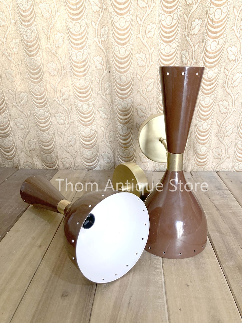 May include: Two brown mid-century modern sconces with brass accents. The sconces have a conical shape with a white interior and small circular perforations. The sconces are on a wooden surface with a patterned background.