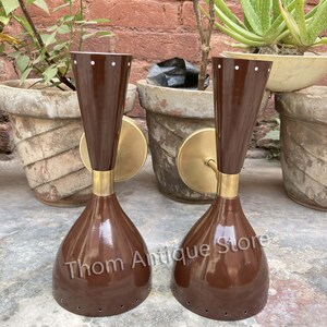 May include: Two vintage brown wall sconces with a brass finish. The sconces have a conical shape with a flared base and a cylindrical top. The sconces are mounted on a round brass plate. The text "Thom Antique Store" is visible on the sconces.