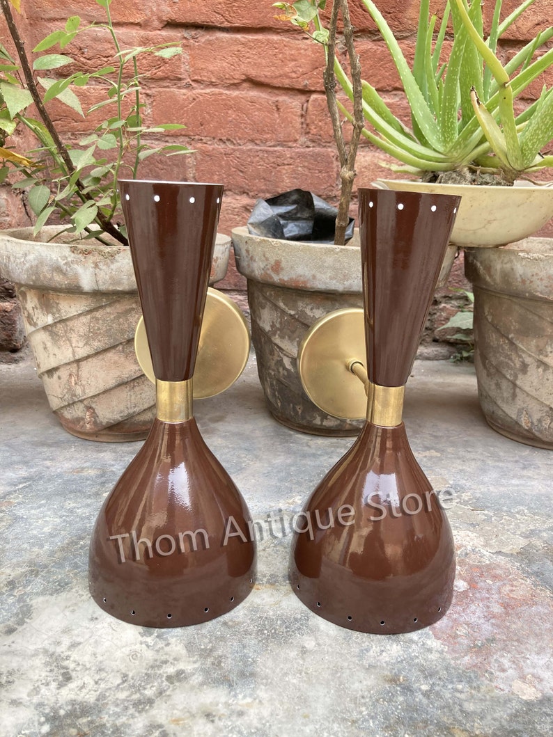 May include: Two vintage brown wall sconces with a brass finish. The sconces have a conical shape with a flared base and a cylindrical top. The sconces are mounted on a round brass plate. The text "Thom Antique Store" is visible on the sconces.