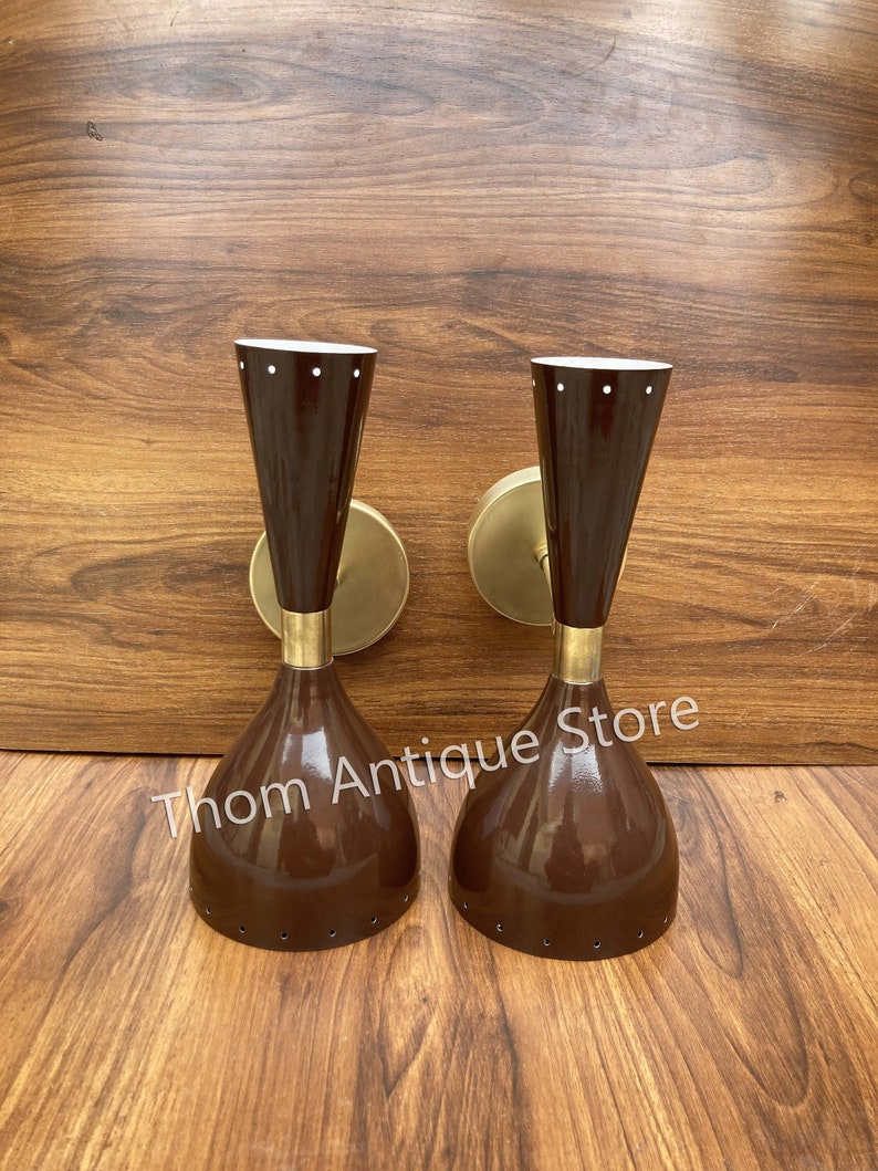 May include: Pair of mid-century modern wall sconces. Each sconce features a brown, conical shade with white dots, a brass neck, and a rounded brass wall mount. The sconces are displayed on a wooden surface. The text "Thom Antique Store" is visible.