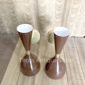 May include: Two vintage brown and brass sconces with a conical shape and white interiors. The sconces have a mid-century modern design, with a round base and a flared top. The text "Thom Antique Store" is visible.