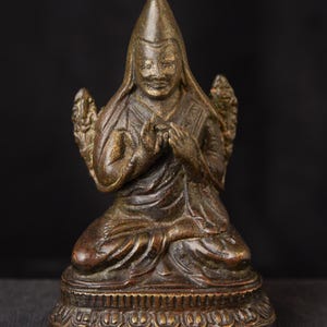 Puede incluir: Estatua de bronce de una figura sentada en posición meditativa. La figura lleva un sombrero puntiagudo y túnicas ornamentadas, con las manos juntas en oración. La estatua tiene rasgos faciales detallados y está montada sobre una base decorativa.
