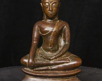 May include: A bronze Buddha statue in a seated meditation pose. The figure has a serene expression, with a detailed headdress and draped robes. The statue is on a decorative base, with a dark background.
