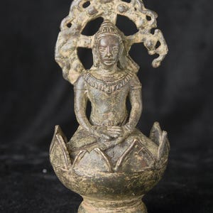 Peut inclure: Une sculpture en métal de couleur bronze foncé représentant une figure assise, les mains posées sur les genoux. La figure est placée dans une base en forme de fleur de lotus stylisée et présente une auréole circulaire ajourée et ornée.