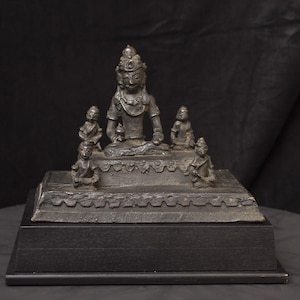May include: A bronze statue of a seated deity with multiple smaller figures. The central figure is adorned with a crown and jewelry. The statue is mounted on a rectangular base with a black wooden stand, showcasing intricate details and craftsmanship.